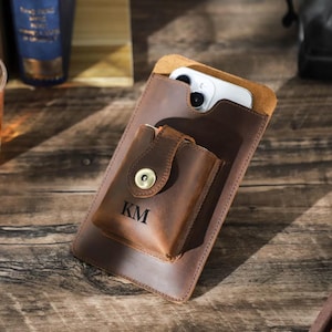 May include: A brown leather phone case with a flap that closes with a snap. The case has a smaller pocket on the front with the initials "KM" embossed on it.