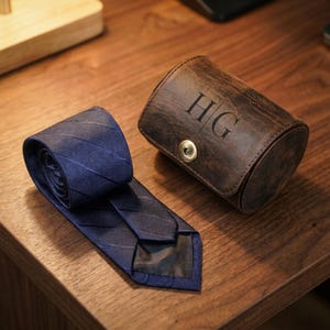 May include: A dark blue necktie with a subtle diagonal stripe pattern alongside a cylindrical brown leather case featuring the embossed letters HG and a single brass-colored metal snap button closure on the front flap.