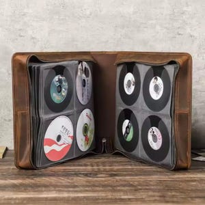 May include: A brown leather CD binder with a zipper closure. The binder is open and has multiple slots for CDs. The CDs are visible and have various designs.