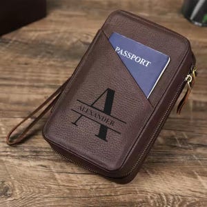 Könnte beinhalten: Eine braune Reisegeldbörse aus Leder mit Reißverschluss. Die Geldbörse hat ein Fach für einen Reisepass und ist mit dem Namen "Alexander" und dem Buchstaben "A" personalisiert.