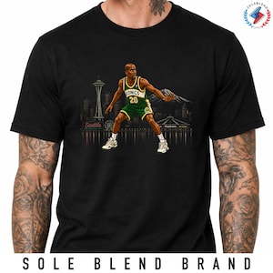 May include: A black t-shirt featuring a graphic of a basketball player in a green and yellow Sonics jersey with the number 20. The background shows a Seattle skyline silhouette with the Space Needle, Mount Rainier, and the text Seattle.