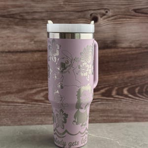 May include: A purple stainless steel travel mug with a white lid. The mug has a floral design with Stitch and other characters from Lilo & Stitch. The text "Nobody gets left behind" is printed on the bottom of the mug.
