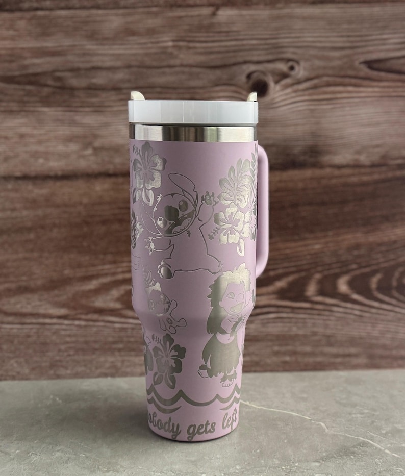 May include: A purple stainless steel travel mug with a white lid. The mug has a floral design with Stitch and other characters from Lilo & Stitch. The text "Nobody gets left behind" is printed on the bottom of the mug.
