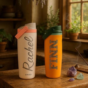 K&ouml;nnte beinhalten: Zwei isolierte Wasserflaschen auf einem Holztisch. Eine ist cremefarben mit dem Namen "Rachel" in silberner Schrift, die andere ist orange mit "FINN" in silbernen Blockbuchstaben. Beide haben Griffe und Deckel. Der Markenname "owala" ist auf der cremefarbenen Flasche aufgedruckt.