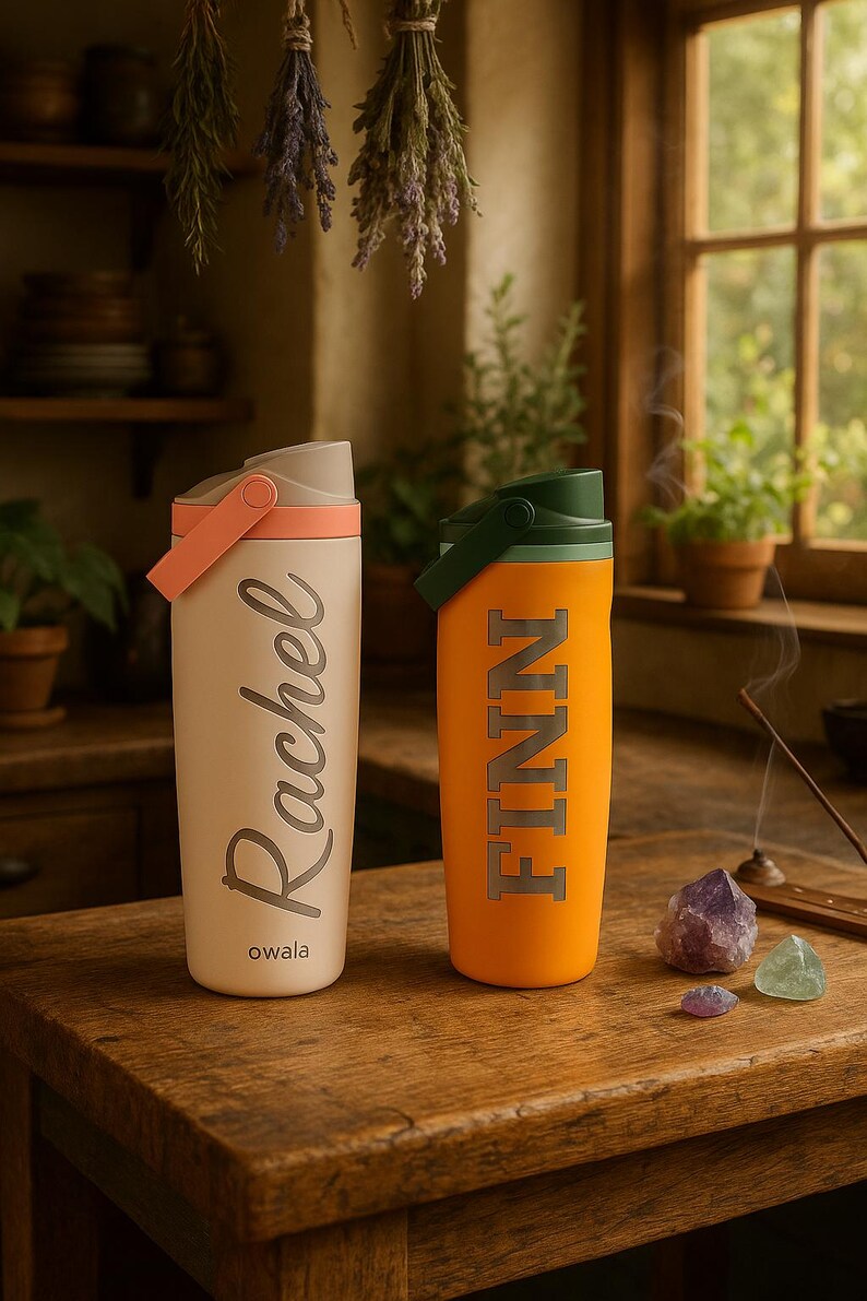 K&ouml;nnte beinhalten: Zwei isolierte Wasserflaschen auf einem Holztisch. Eine ist cremefarben mit dem Namen "Rachel" in silberner Schrift, die andere ist orange mit "FINN" in silbernen Blockbuchstaben. Beide haben Griffe und Deckel. Der Markenname "owala" ist auf der cremefarbenen Flasche aufgedruckt.