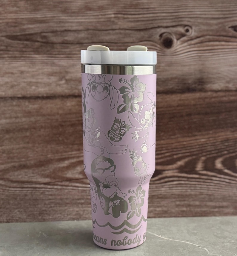 May include: A lavender stainless steel tumbler with a white lid and a silver design of Stitch and other characters from Lilo & Stitch. The tumbler has the text "Ohana means nobody gets left behind" at the bottom.
