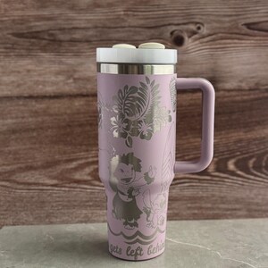 May include: A lavender stainless steel travel mug with a white lid and a handle. The mug has a design of Stitch and Lilo from the Disney movie Lilo & Stitch. The design is engraved on the mug and includes tropical flowers and leaves. The text "gets left behind" is at the bottom of the mug.