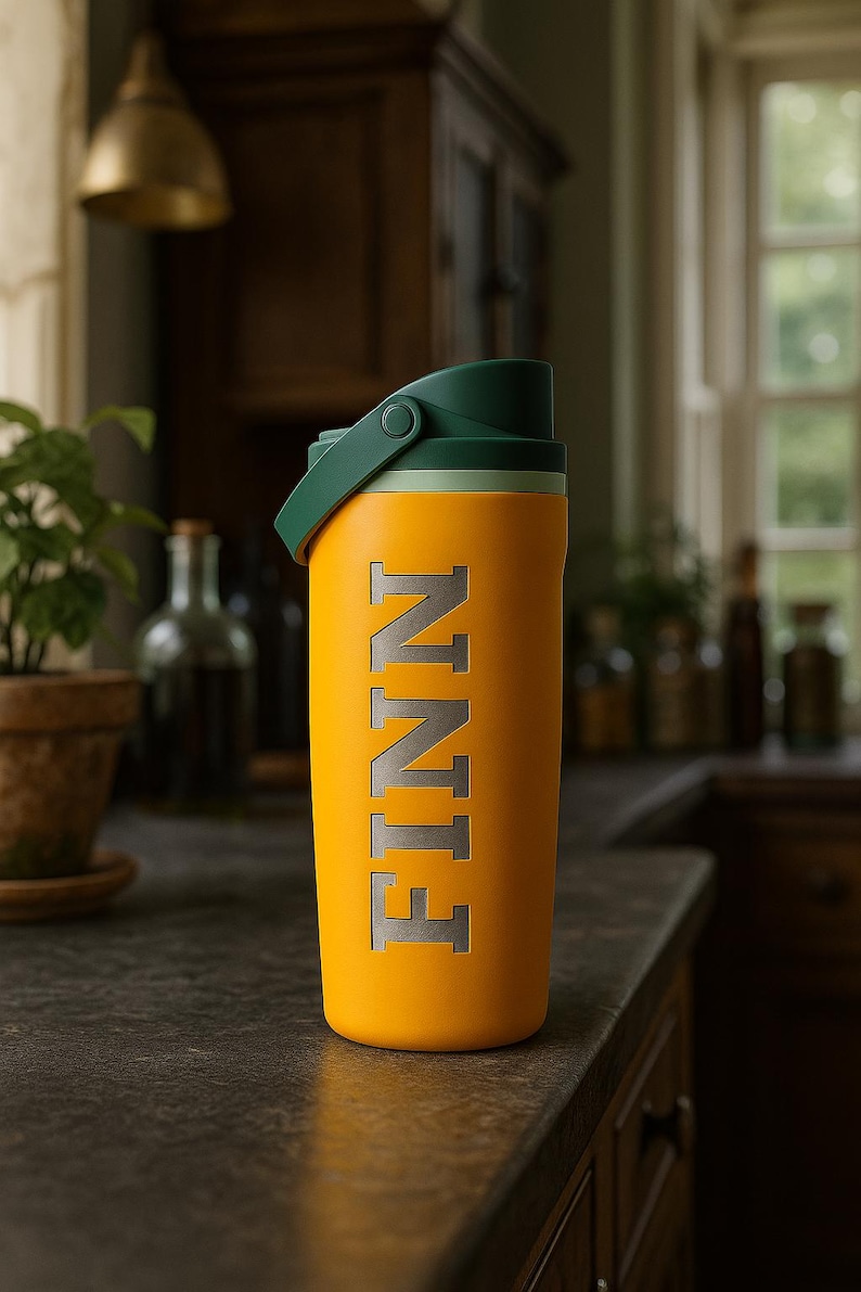 K&ouml;nnte beinhalten: Eine gelbe Wasserflasche mit dem eingravierten Namen "FINN". Die Flasche hat einen gr&uuml;nen Deckel und Griff. Die Flasche ist zylindrisch und scheint aus Metall zu sein. Der Hintergrund ist eine K&uuml;chenszene.