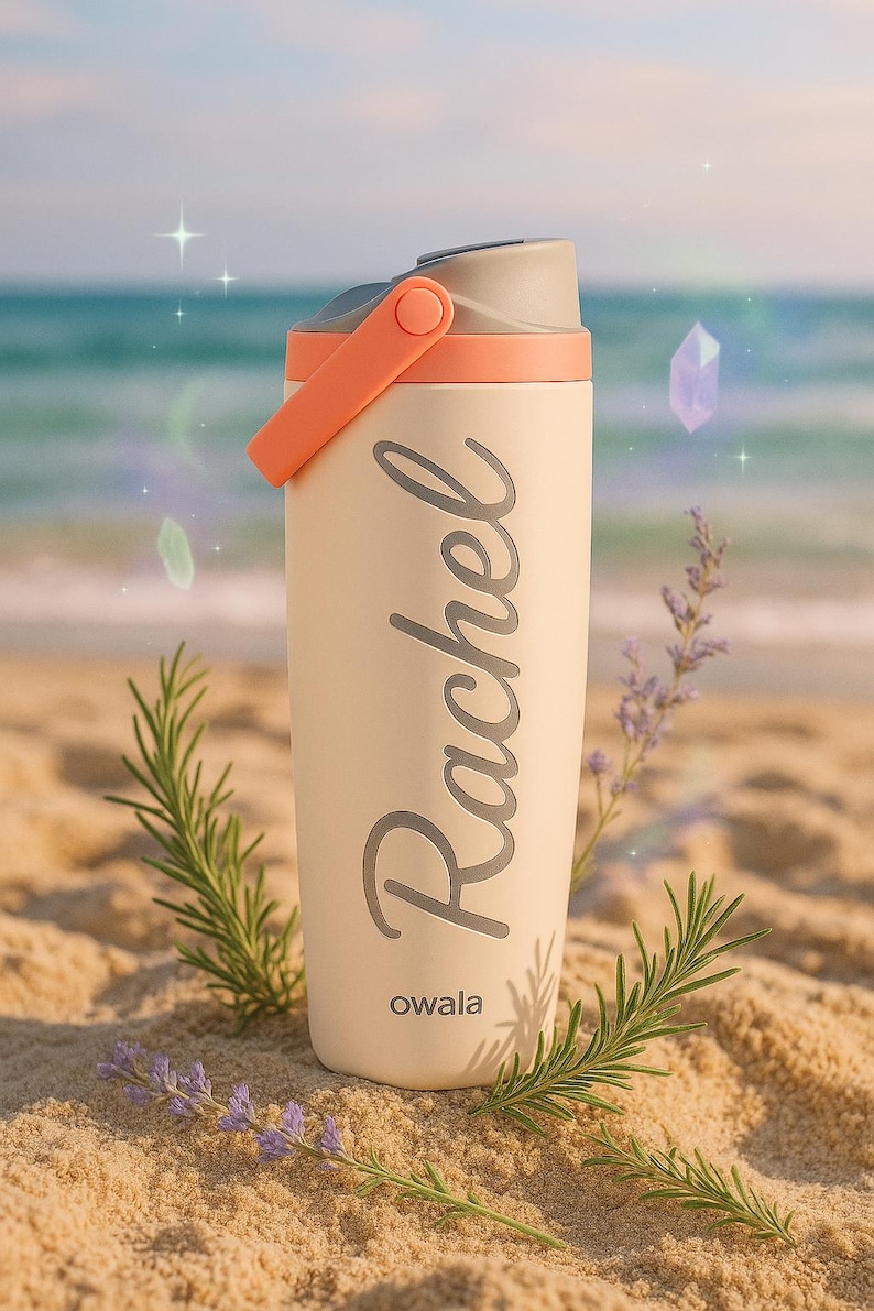 K&ouml;nnte beinhalten: Eine cremefarbene Owala-Trinkflasche mit dem Namen "Rachel" in Schreibschrift. Die Flasche hat einen grauen Deckel und einen orangefarbenen Griff. Der Hintergrund zeigt eine Strandszene mit Sand, Wasser und Gr&uuml;n.