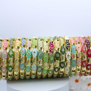May include: A collection of gold-toned bangles, each adorned with colorful floral designs. The bangles feature various colors, including pink, green, blue, and white, with a small crystal accent in the center of each flower. These bracelets are a vibrant accessory.