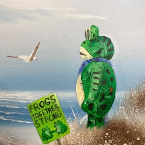 May include: An oil painting depicts a green frog figure on a beach, with a sign that reads "Frogs Together Strong." Two seagulls fly in the sky above the ocean. The frog has black spots and a blue scarf.