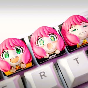 Peut inclure: Gros plan sur trois touches de clavier personnalisées représentant un personnage d'anime aux cheveux roses et aux yeux verts. Les touches sont sur un clavier blanc avec d'autres touches visibles. Les touches ont des expressions faciales différentes.