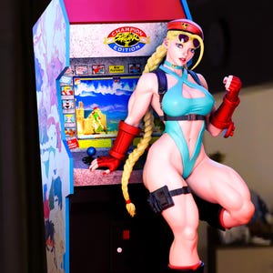 以下が含まれることがあります： ストリートファイターIIの女性キャラクターのフィギュア。水色のボディスーツ、赤い手袋、赤い帽子を着用しています。キャラクターは、チャンピオンストリートファイターIIのアーケードキャビネットの隣にポーズをとっています。