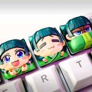 Puede incluir: Tres teclas personalizadas con diseños de personajes de estilo anime. Cada tecla tiene una expresión facial diferente y es principalmente verde, beige y azul. Las teclas están en un teclado blanco con letras negras.