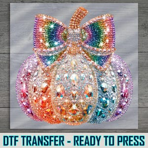 May include: A colorful pumpkin design with a bow, covered in sparkling rhinestones. The pumpkin is in shades of orange, pink, purple, and blue. The text "DTF TRANSFER - READY TO PRESS" is at the bottom.