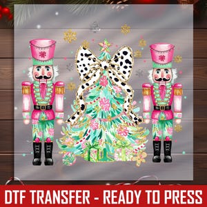 May include: A festive Christmas-themed design featuring two nutcrackers flanking a decorated Christmas tree. The tree is adorned with a dalmatian print bow and gold accents. The nutcrackers wear pink and green uniforms. The image includes the text "DTF TRANSFER - READY TO PRESS".