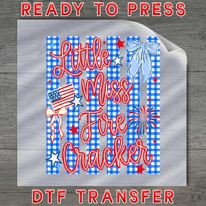 May include: A DTF transfer sheet featuring a blue and white gingham striped background with the red text Little Miss Fire Cracker. The design includes a small American flag, a blue bow, a red and blue firework burst, and several red and white stars.