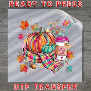 May include: A DTF transfer sheet featuring a colorful illustration of a large orange pumpkin, a smaller teal pumpkin, and a coffee cup with a pink lid. The cup has the text hello Fall. A plaid scarf, autumn leaves, and an acorn surround the items.