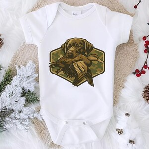 A white baby onesie with a graphic of a dog holding a bird is displayed on a white surface, surrounded by pine cones and red berries.