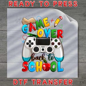 May include: A DTF transfer sheet featuring a colorful graphic with a video game controller at the center. The text reads GAME OVER back to SCHOOL. Surrounding elements include an open book, a yellow pencil, a green crayon, and a red apple.