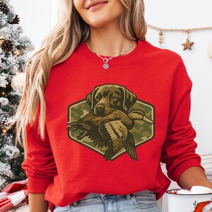 A woman wearing a red sweater with a graphic of a hunting dog holding a duck, standing in front of a Christmas tree.