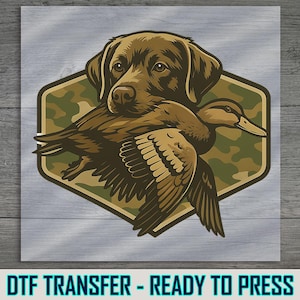May include: A brown Labrador Retriever dog with a duck in its mouth, set against a camouflage background within a hexagonal shape. The words "DTF TRANSFER - READY TO PRESS" are at the bottom of the image.