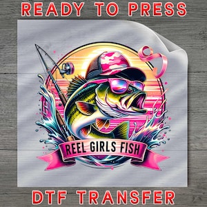 May include: A colorful graphic featuring a fish wearing sunglasses and a cap, with a fishing rod in the background. The text "REEL GIRLS FISH" is on a banner. The words "READY TO PRESS" and "DTF TRANSFER" are also visible.