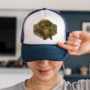 A woman is holding up a blue and white baseball cap with a hunting dog on it, smiling at the camera.