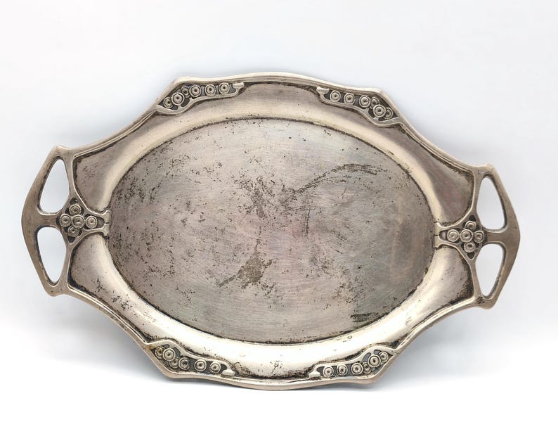 Puede incluir: Una bandeja de servir antigua de color plateado con un centro ovalado y asas decorativas. La bandeja tiene detalles ornamentados alrededor de los bordes y las asas, con un acabado ligeramente desgastado. Una pieza vintage para servir o exhibir.