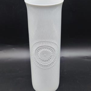 May include: A tall, white ceramic vase with a textured, vertical line pattern. The vase features a circular, embossed design near the center. The vase has a flared top and a smooth, rounded base. The vase is approximately 12 inches tall.