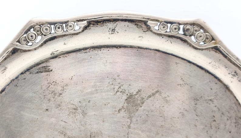 Puede incluir: Primer plano de un plato de plata deslustrado con un borde ornamental y decorativo. El plato tiene una forma ligeramente octogonal con una superficie lisa y reflectante. El borde presenta un patr&oacute;n repetitivo de peque&ntilde;os c&iacute;rculos.