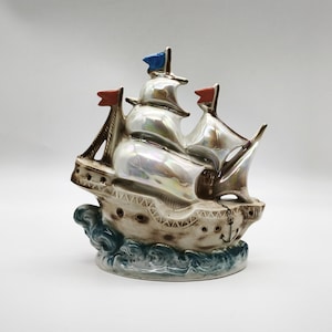 Könnte beinhalten: Keramikschiffsfigur mit schillernden Segeln und drei roten und blauen Flaggen. Das Schiff hat einen detaillierten Rumpf mit einem dekorativen Muster und steht auf einem Sockel, der Meereswellen darstellt. Ein Anker ist sichtbar.