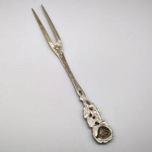May include: Silver-toned two-pronged fork with floral and leaf detailing. The handle features an engraved design and a decorative end with a rose-like emblem. A vintage utensil for serving or display.
