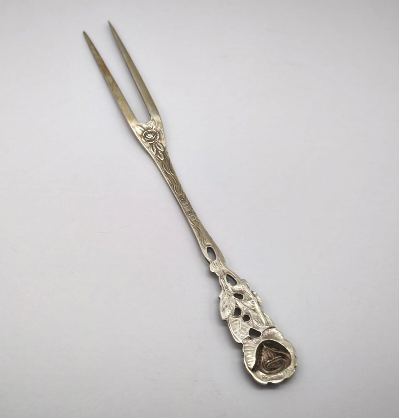 May include: Silver-toned two-pronged fork with floral and leaf detailing. The handle features an engraved design and a decorative end with a rose-like emblem. A vintage utensil for serving or display.