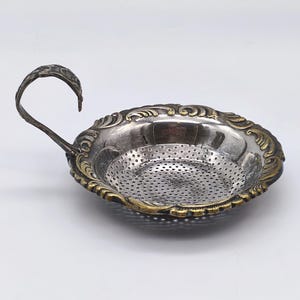 Op de afbeelding: Een zilveren thee-ei met een sierlijke handgreep en een decoratieve goudkleurige rand. Het thee-ei heeft een geperforeerde kom voor het filteren van theebladeren. De handgreep buigt omhoog en de rand heeft een gedetailleerd, reliëf ontwerp.