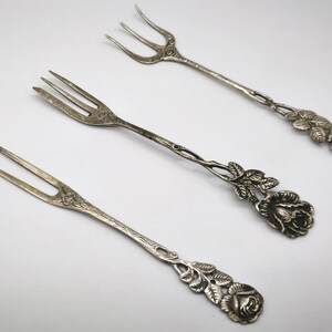 May include: Three antique silver forks with a floral rose design. The forks have two or three tines and decorative rose and leaf accents on the handles. The forks are arranged on a white surface.
