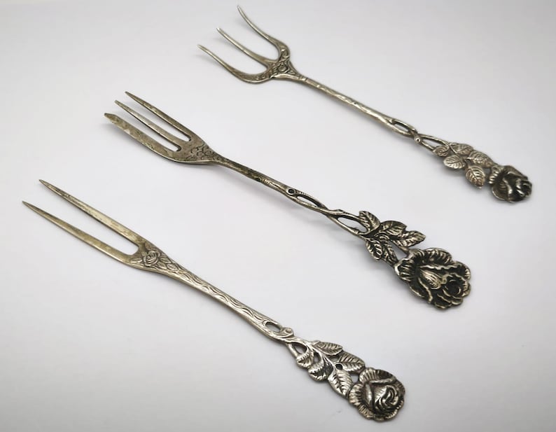 May include: Three antique silver forks with a floral rose design. The forks have two or three tines and decorative rose and leaf accents on the handles. The forks are arranged on a white surface.