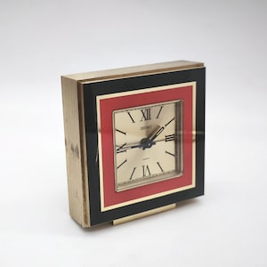 Puede incluir: Un reloj de cuarzo Seiko vintage con caja dorada. La esfera es dorada con manecillas negras y números romanos. La esfera está enmarcada por un borde rojo y negro. El reloj mide aproximadamente 7.6 cm de ancho.