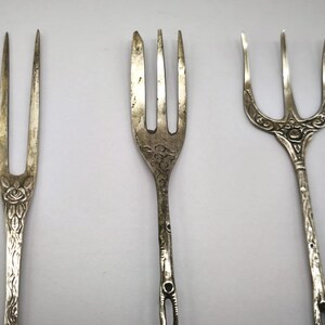 May include: Three antique silver forks with ornate designs. The forks have different styles, including floral and etched patterns. The tines vary in number and shape, showcasing the craftsmanship of each piece. The forks are displayed against a plain background.
