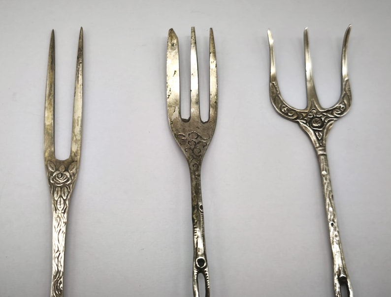 May include: Three antique silver forks with ornate designs. The forks have different styles, including floral and etched patterns. The tines vary in number and shape, showcasing the craftsmanship of each piece. The forks are displayed against a plain background.