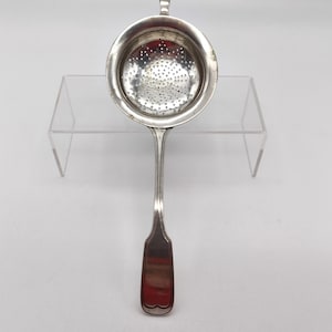 Op de afbeelding: Een zilveren thee-ei met een ronde kom met talrijke kleine gaatjes. Het heeft een lange, slanke steel met een decoratief uiteinde en een kleine lus aan de rand.