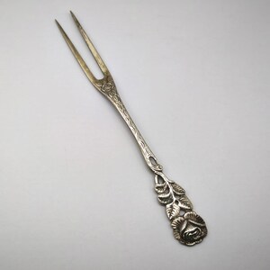 May include: Silver-toned serving fork with two tines. The handle features a detailed floral design, including a rose and leaves. The fork is likely made of metal and is designed for serving food.