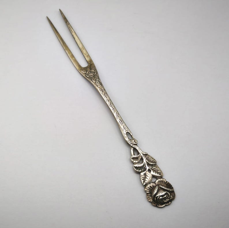 May include: Silver-toned serving fork with two tines. The handle features a detailed floral design, including a rose and leaves. The fork is likely made of metal and is designed for serving food.