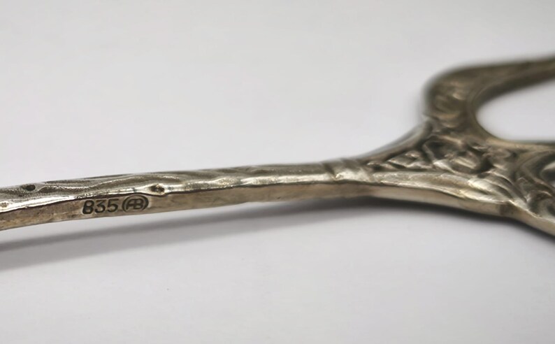 May include: Close-up of a silver serving utensil with an ornate, floral design. The handle has a textured surface and the hallmark "835" is visible. The utensil is likely vintage and used for serving food.