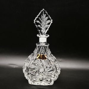 May include: A clear crystal decanter with a leaf-shaped stopper. The decanter has a detailed, cut-glass design with a starburst pattern on the base. A gold label is visible on the front of the decanter. The background is black.