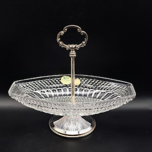 Op de afbeelding: Een glazen serveerschaal met een decoratief, geslepen glaspatroon. De schaal heeft een zilverkleurige voet en een zilverkleurige handgreep.