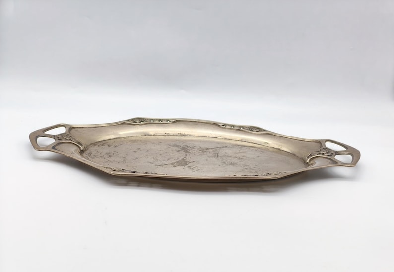 Puede incluir: Una bandeja de servir plateada, de estilo Art Nouveau. La bandeja ovalada alargada tiene asas decorativas con dise&ntilde;os calados. El borde presenta un borde festoneado sutil. La superficie de la bandeja muestra signos de edad y uso.