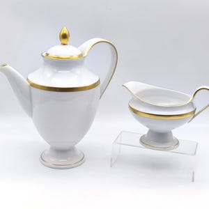 May include: A white porcelain tea set with gold trim. The set includes a teapot with a lid and a creamer. The teapot has a curved handle and spout. The creamer sits on a pedestal and has a curved handle.
