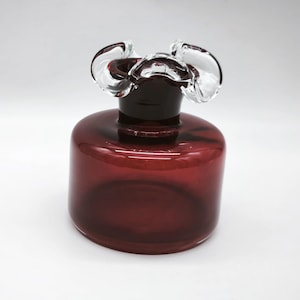 Könnte beinhalten: Eine bordeauxfarbene Glasvase mit einem klaren, gerüschten Oberteil. Die Vase hat einen zylindrischen Körper und einen dunklen Hals. Die transparente Oberseite hat eine blütenartige Form. Die Vase wird wahrscheinlich zu Dekorationszwecken verwendet.