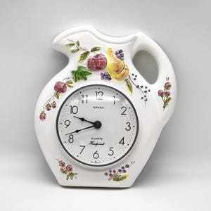 May include: White ceramic pitcher-shaped wall clock with a black clock face. The clock features a fruit and floral design with a handle. The clock face has black numbers and hands. The clock is marked "Kienzle" and "Quartz Handcrafted".